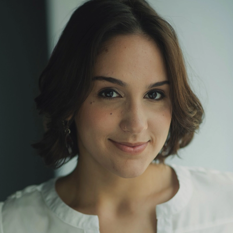 Kyra Paternoster, a young woman with short brown hair and light skin, smiles softly at the camera. She wears a white blouse with a round neckline and subtle earrings, her face highlighted against a softly blurred background of dark and light tones.