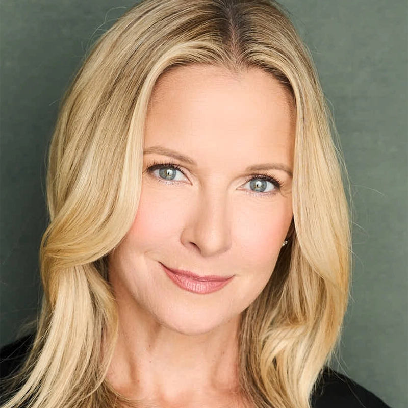 Meredith Patterson, with long, straight blonde hair and blue eyes, smiles softly. She is wearing a black top against a muted green background. Bright lighting highlights her clear skin and natural makeup.