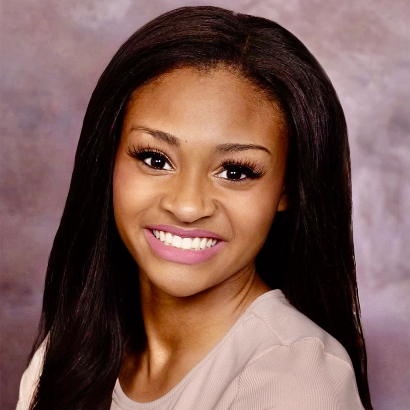 Madelyn Primer, a young woman with long, straight dark hair and brown skin, smiles at the camera. She wears a beige top and natural makeup with pink lipstick against a softly blurred purple and gray background.