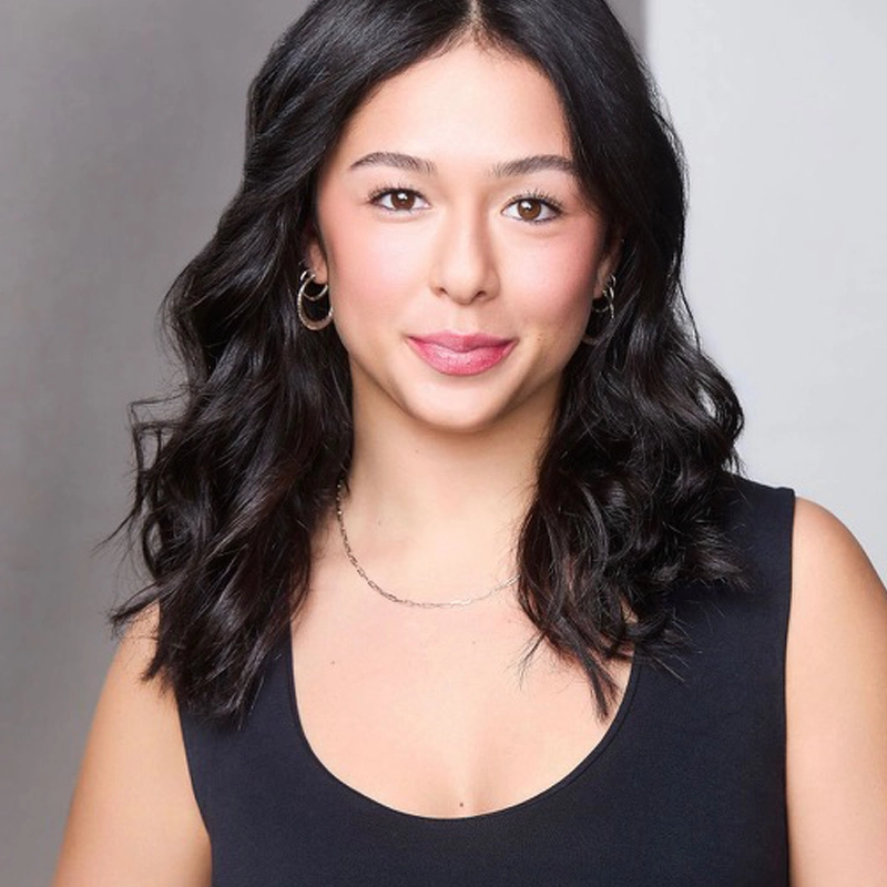 Sloane Ptashek, a young woman with wavy, shoulder-length dark hair, wearing a sleeveless black top and small hoop earrings, smiles softly at the camera against a neutral gray background. She has a light complexion and natural makeup.