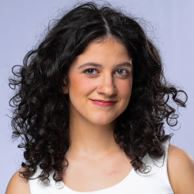 A young woman with medium skin and dark, curly hair smiles gently at the camera. She has a small nose piercing, wears natural makeup, and is dressed in a white sleeveless top. The background is a soft, light purple.