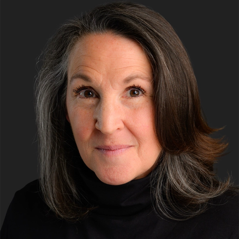 Marianna Raho, a middle-aged woman with long, wavy gray and brown hair, wears a black turtleneck and gazes warmly into the camera. Her gentle, confident expression is highlighted by soft lighting against a solid black background.