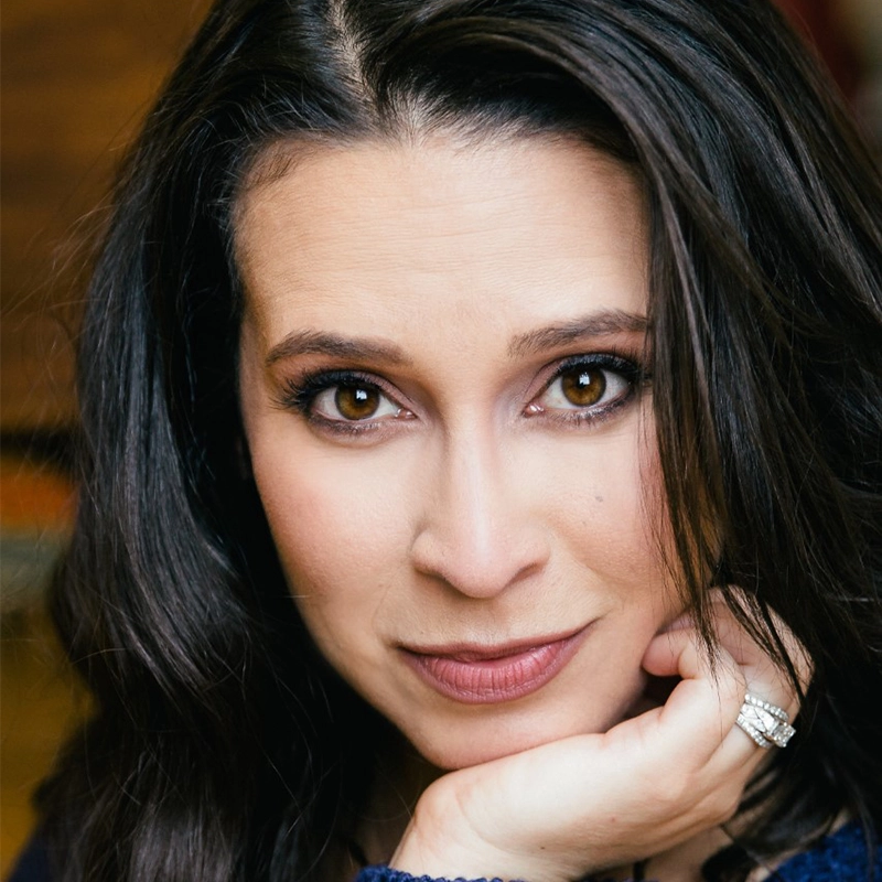 A woman with long, dark brown hair, brown eyes, and defined eyebrows looks directly at the camera. She rests her chin on her hand, showing a silver ring on her finger. She wears light makeup and a dark top; the background is softly blurred.