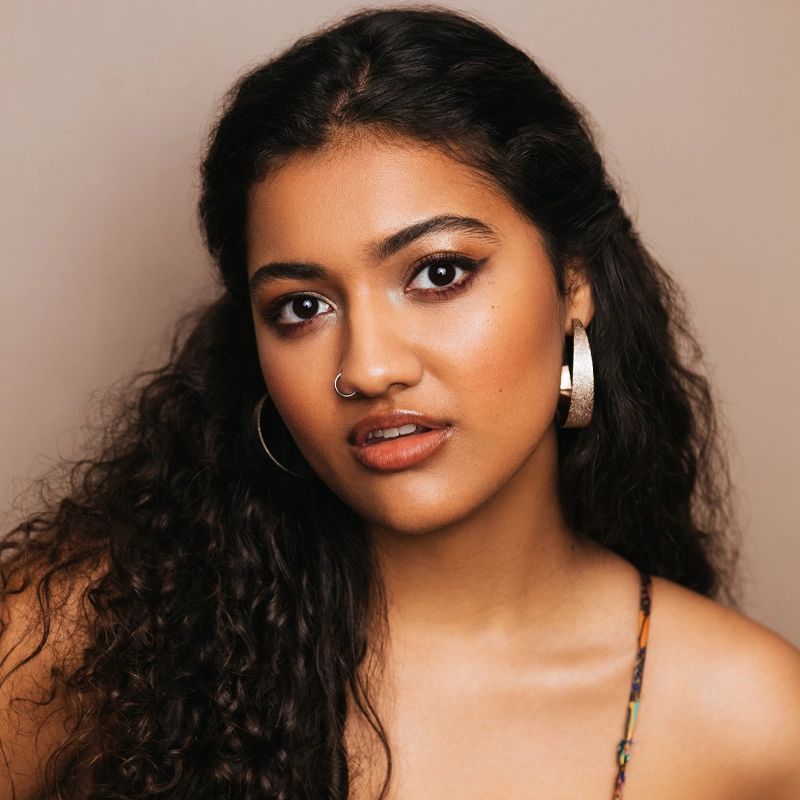 A young woman with long, curly dark hair looks at the camera. She wears large hoop earrings, a nose ring, and subtle makeup with winged eyeliner and nude lipstick. The background is plain beige, and she has a confident, calm expression.