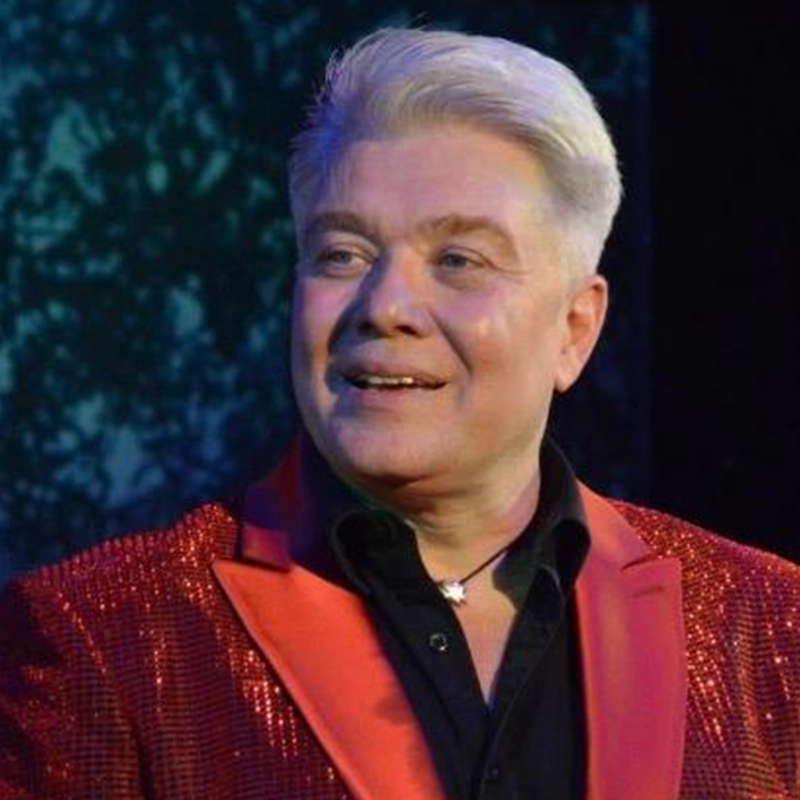 A smiling man with short, white hair—Edmund Bagnell—wears a sparkling red blazer with a black lapel over a black shirt. He stands against a dark, blurred background, illuminated by dramatic stage lighting.