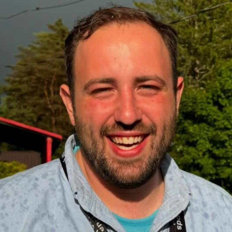 Dominique Alvarez, a smiling man with short brown hair and a beard, stands outdoors in a light blue shirt and black lanyard. He looks happy and sunlit, with trees and a red-roofed building under a partly cloudy sky behind him.