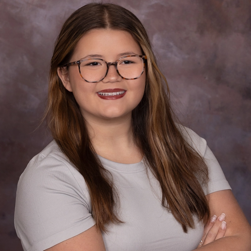 Chloe Robertson, a young woman with long brown hair and glasses, smiles at the camera. She wears a light gray short-sleeve top with her arms crossed, set against a blurred backdrop of brown and purple tones for a studio portrait feel.