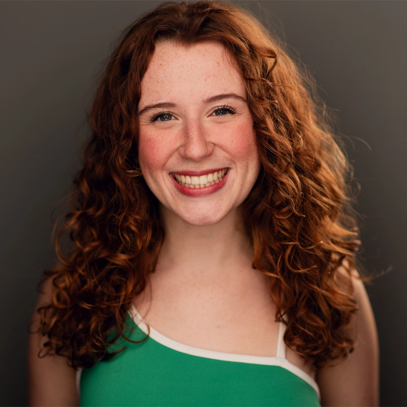 A young woman, resembling Heléne Rosborough, with long, curly red hair and fair skin smiles widely at the camera. She has freckles on her cheeks and is wearing a green tank top with white trim. The plain gray background keeps her as the focal point.