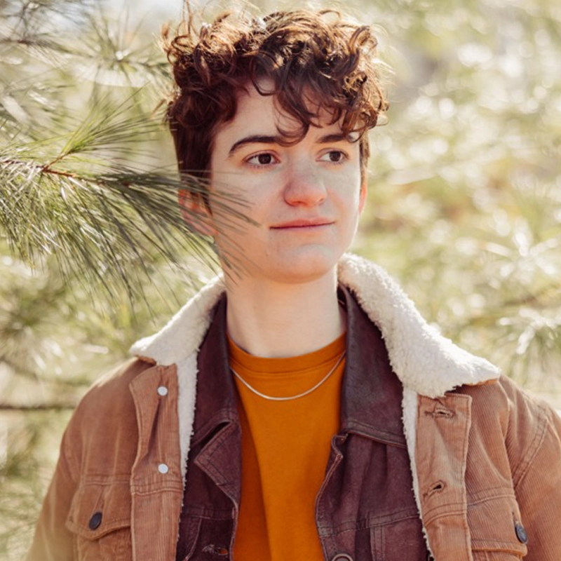 Talia Rubenstein, with short, curly brown hair, stands among pine trees in a brown corduroy jacket with a white sherpa collar. Sunlight softly highlights her face as she gazes slightly to the right, wearing an orange shirt and a thin necklace.
