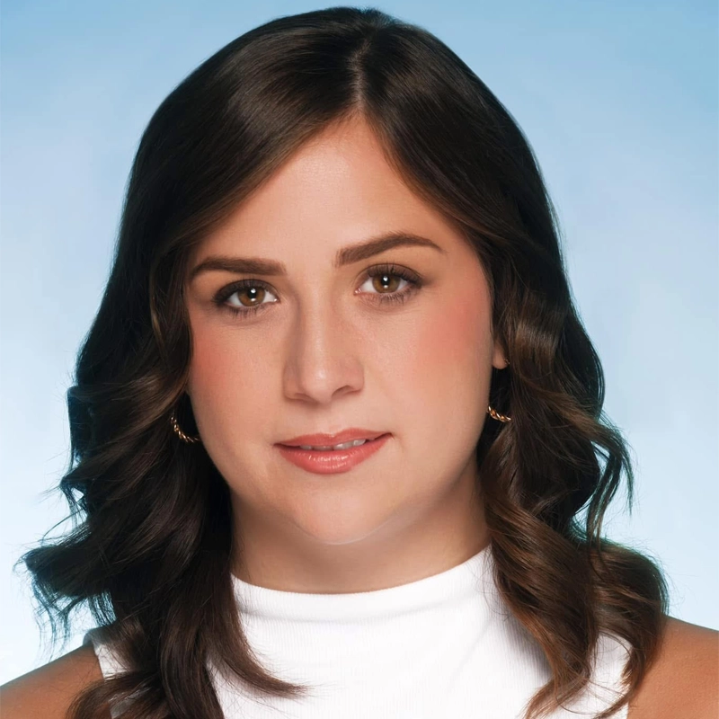 A woman with medium-length, wavy brown hair and brown eyes is facing the camera against a light blue background. She is wearing a sleeveless white top and small hoop earrings, with a neutral expression and natural makeup.