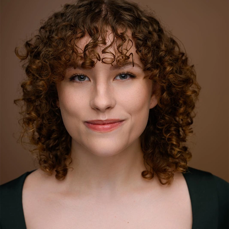 Jordyn Rubinsky, a young woman with fair skin and curly brown hair, smiles softly at the camera. She has blue eyes, light makeup, and wears a dark top. The solid warm brown background creates a professional and pleasant portrait.