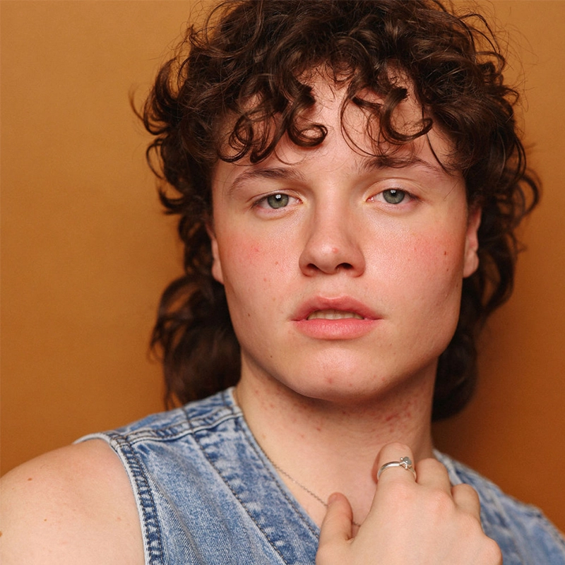A young person with curly brown hair and fair skin gazes at the camera with a neutral expression. Taylor Leccese wears a sleeveless denim vest and a silver ring on their finger, set against a warm brown background.