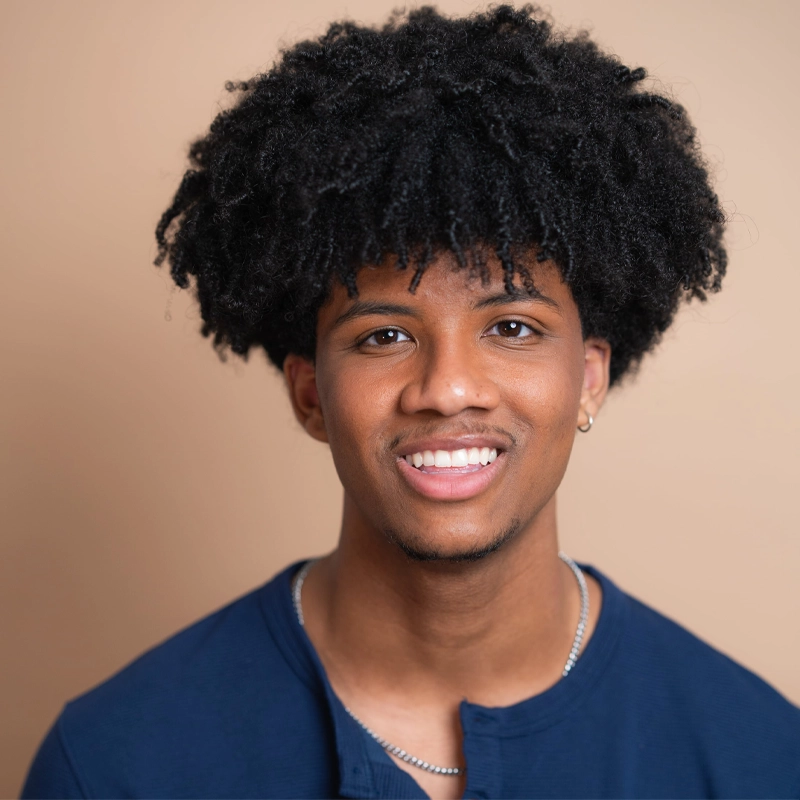 A young man with medium brown skin, short beard, and voluminous curly black hair smiles at the camera. Garan Salandy wears a blue shirt, silver necklaces, and a small hoop earring in one ear. The background is plain beige.