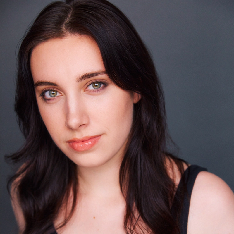 A young woman with long, dark brown hair and light skin looks at the camera with a slight smile. She has green eyes, subtle makeup, and wears a black top. The portrait is in the style of photographer Michael Raul Perez against a plain, dark gray background.