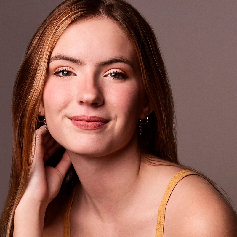 A young woman with long, light brown hair smiles softly. She wears natural makeup, small hoop earrings, and a mustard-yellow tank top. Her hand gently touches her neck as she poses against a neutral, light brown background.