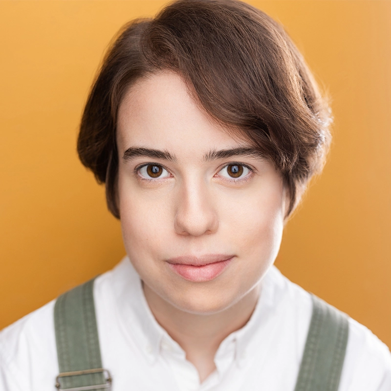 A person with short brown hair and fair skin faces the camera, wearing a white shirt with green suspenders. Their expression is neutral, set against a bright orange background with soft, even lighting.