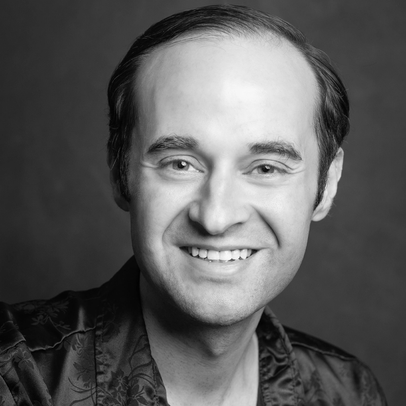 A black and white portrait of Patrick Schaefer, a smiling man with short, dark hair, wearing a patterned shirt. He has light skin and is facing forward against a plain, dark background. The lighting highlights his friendly features.