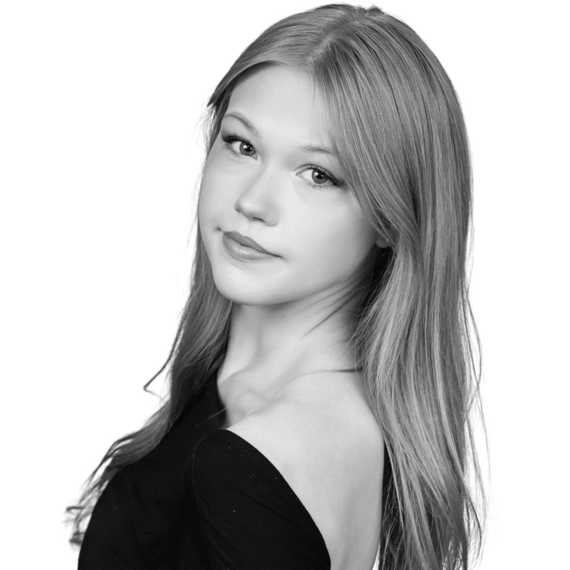 Black and white portrait of Libby Schwartz, a young woman with long, straight hair, looking directly at the camera with a calm expression. She wears a dark, off-the-shoulder top and has a slight, gentle smile against a plain white background.