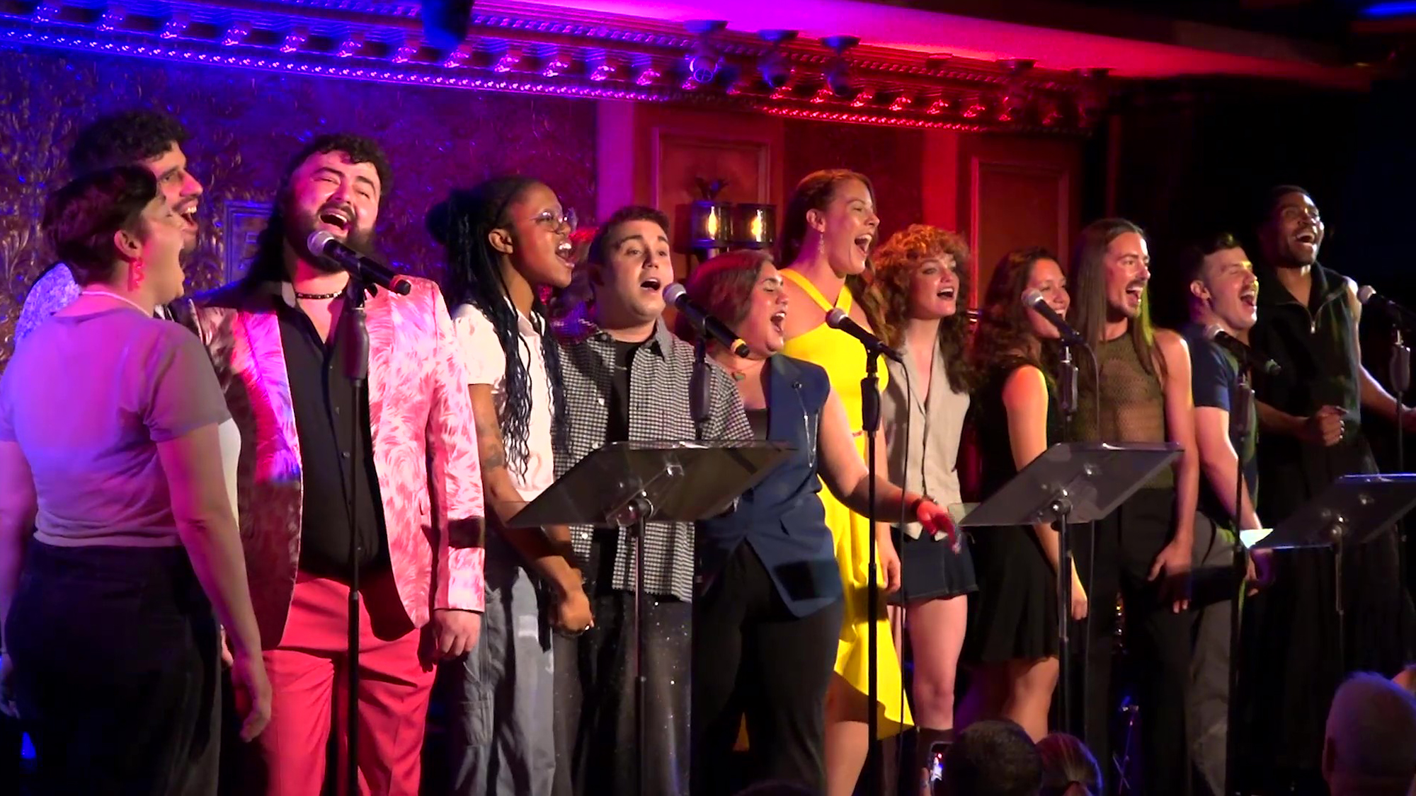 Screencap of Coming Out Correct Size A diverse group of nine singers stands in a row on stage, passionately singing songs from new musicals. They wear colorful outfits under bright purple and pink lights, performing expressively before a warmly lit, ornate backdrop.