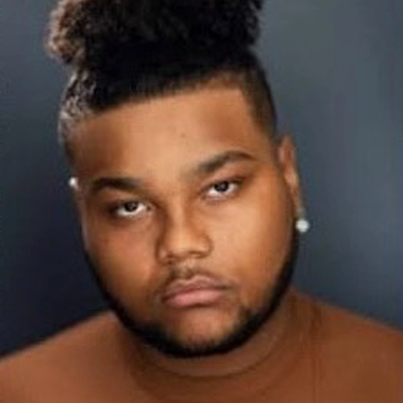 A young man with a medium-dark complexion, short beard, and mustache stares directly at the camera. He has curly hair styled in a high bun and wears a brown shirt and a single stud earring, set against a dark, blurred background.