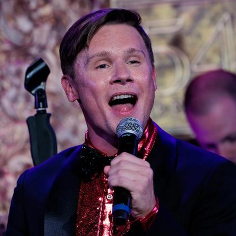 Edmund Bagnell, a man with short brown hair, sings into a microphone. He wears a shiny red shirt, black sequin bow tie, and dark jacket. The blurred background features a gold 
