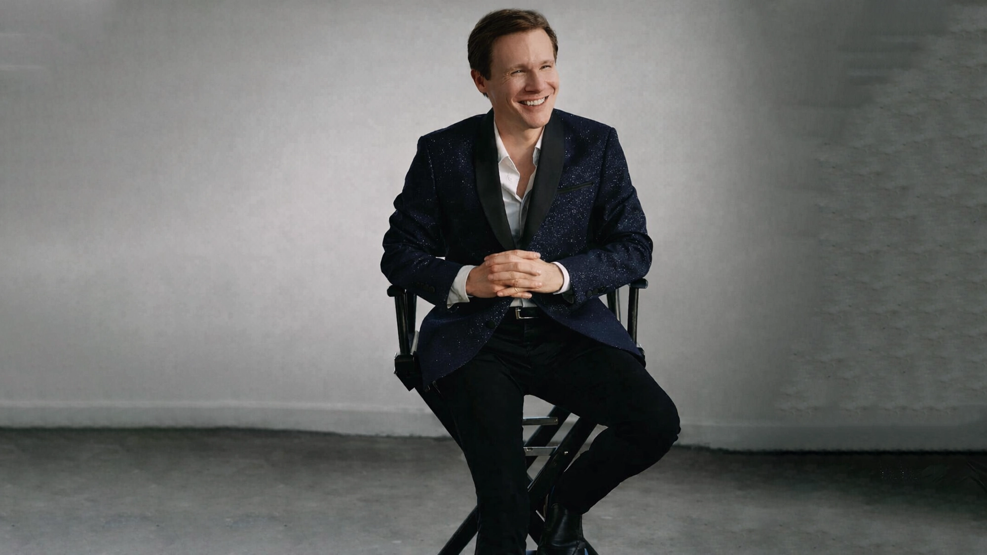A smiling man, Seth Sikes, sits on a director’s chair against a plain, light gray backdrop. He wears a dark, textured suit jacket, white dress shirt, black pants, and black shoes—ready to celebrate Judy Garland's birthday in style.