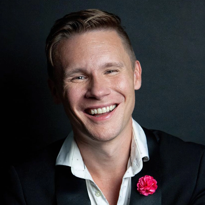 A man with light skin and short, light brown hair smiles broadly. Edmund Bagnell wears a black blazer over a white shirt with a subtle pattern, and a bright pink flower pin on his lapel. The dark background highlights his cheerful expression.
