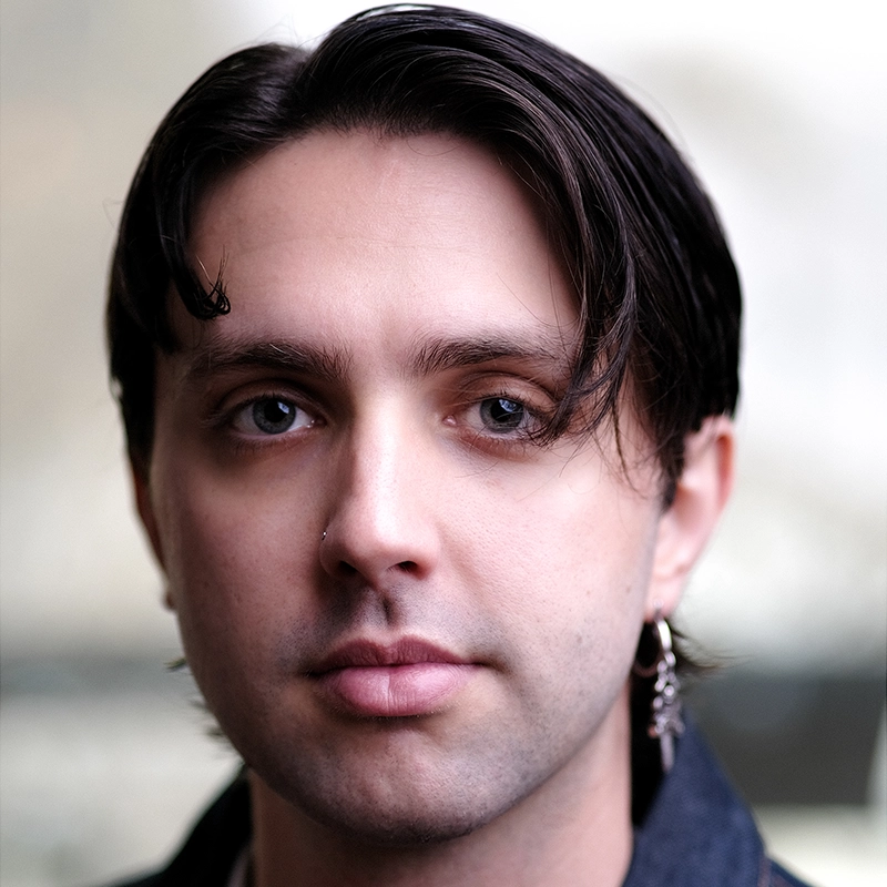 Casey Martin Klein, with light skin and dark brown, straight hair parted down the middle, stares into the camera. A small nose ring graces their left nostril and a dangling earring hangs from their left ear. The background is softly blurred and neutral in color.