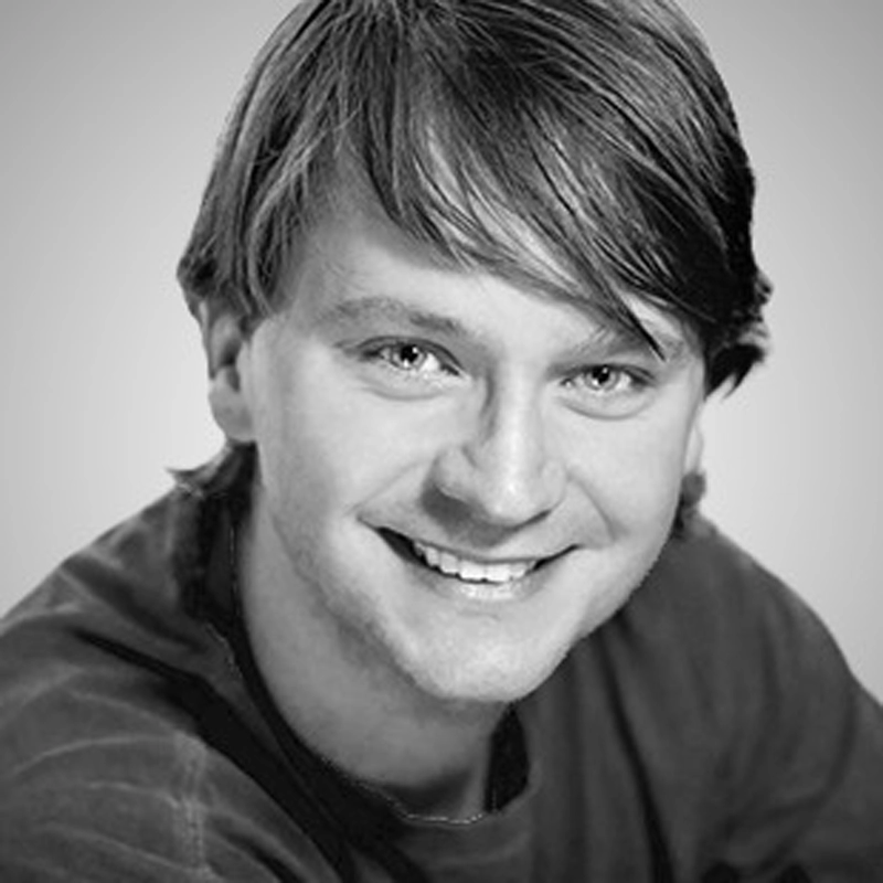 A young man with straight, light brown hair and fair skin smiles at the camera. He is wearing a casual dark shirt. The portrait is black and white with even lighting and a plain, light background.