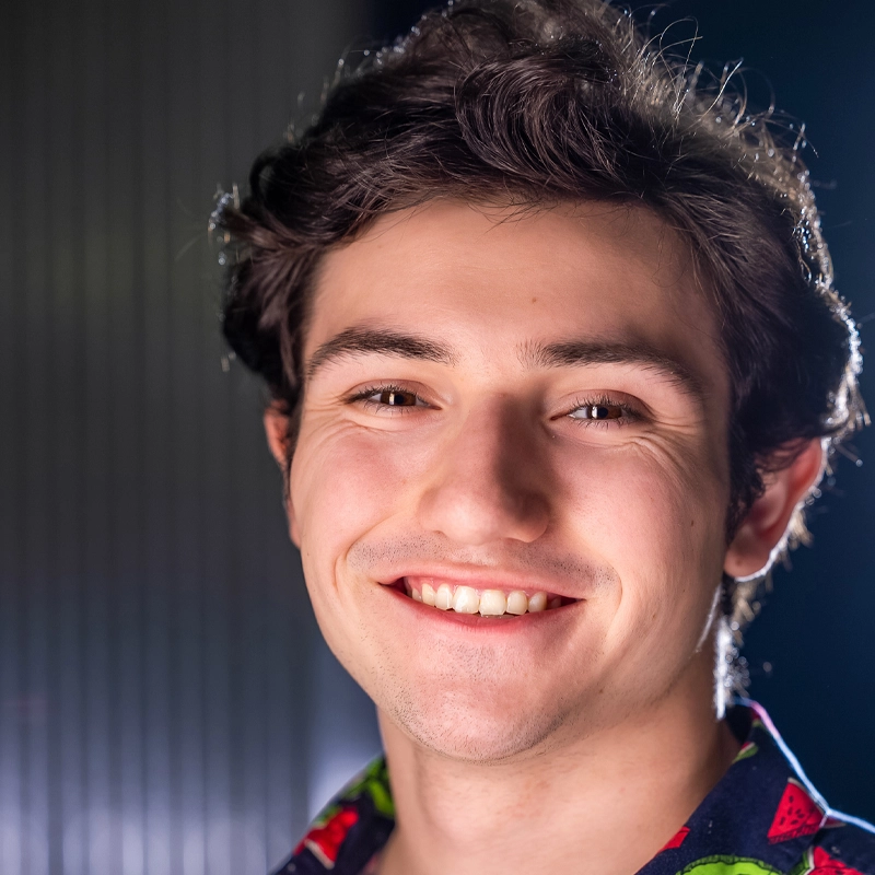 Daniel Sidoni, a young man with wavy dark hair, smiles brightly at the camera. He has light skin, brown eyes, and wears a dark shirt with a colorful pattern. The softly blurred background features subtle vertical lines and gentle highlights on his face.