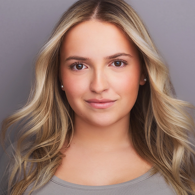Sienna Mello, a young woman with long, wavy blonde hair and fair skin, smiles softly at the camera. She wears a light gray top and stud earrings. The plain, neutral background draws focus to her face and natural makeup.