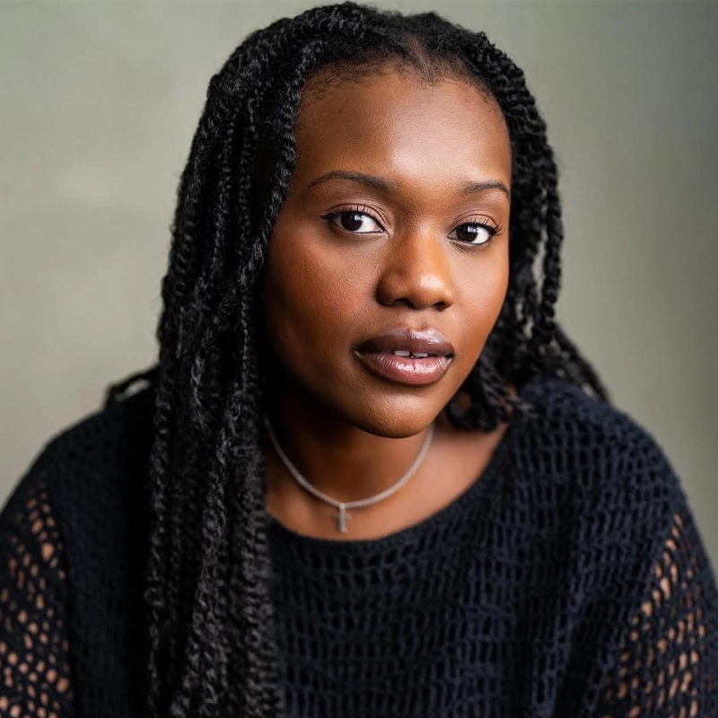 Tenneh Sillah, with long black braided hair and smooth dark skin, looks at the camera in a black textured sweater and a thin necklace. She poses against a light gray background with a calm, confident expression.