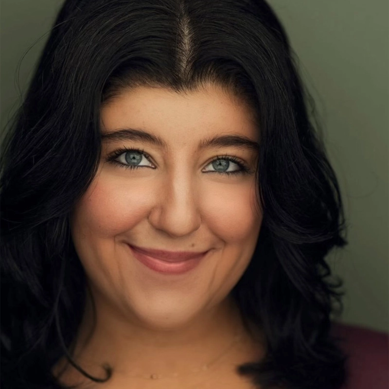 A woman with wavy, shoulder-length dark hair smiles warmly at the camera. She has blue eyes, smooth skin, and is wearing natural makeup. The background is a plain, muted green, and she is dressed in a dark top.