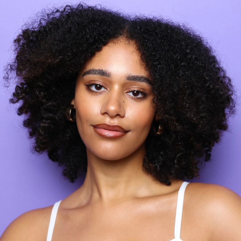 Maya Sistruck, with medium-brown skin and natural, voluminous curly hair, poses against a solid purple background. She wears gold hoop earrings, natural makeup, and a white spaghetti strap top, exuding a calm, confident expression.