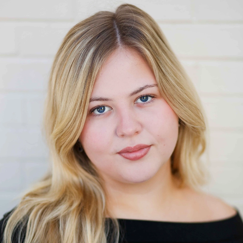 Aubrie Ofner, a young woman with long blonde hair and blue eyes, gazes at the camera with a slight smile. She wears a black off-the-shoulder top against a softly blurred pale brick wall, lit by natural light.