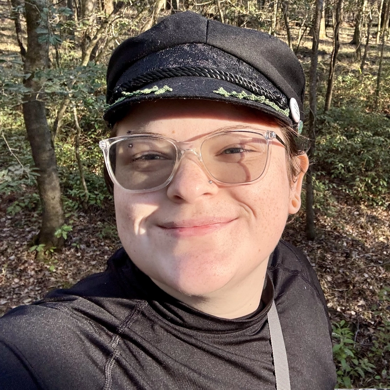 Remy Stevens, with light skin, clear glasses, and a small pierced nose, smiles outdoors. They wear a black cap with decorative trim, a black top, and a gray strap as sunlight filters through trees in the wooded area behind them.