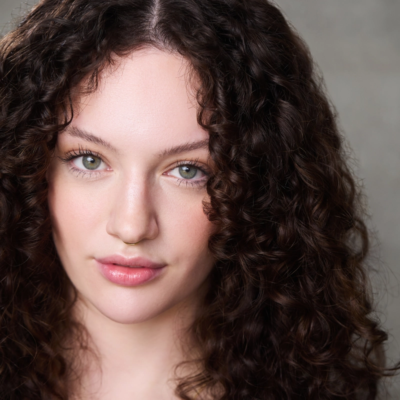 A young woman with fair skin and long, dark, curly hair gazes directly at the camera. She has green eyes, slightly arched eyebrows, and natural makeup with soft pink lips. The background is plain and gray, highlighting her facial features.