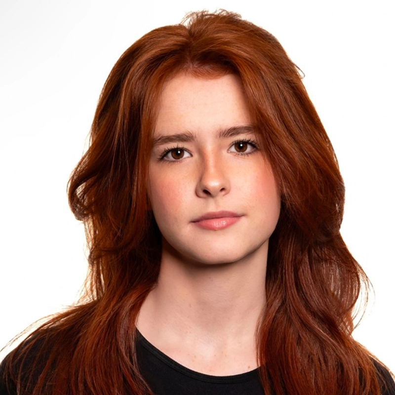 A young person with long, wavy reddish-brown hair and fair skin looks at the camera with a neutral expression. They are wearing a black top, and the background is plain white, giving the image a clean and bright appearance.