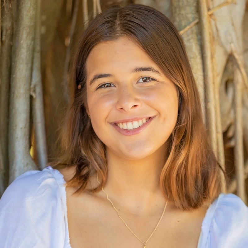 Ava Szlabowicz, a young woman with medium-length brown hair, smiles warmly at the camera. She wears a light blue, puff-sleeve blouse and a delicate gold necklace. Textured tree trunks and soft sunlight enhance her natural features.