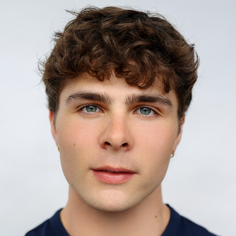 Aubrie Ofner, a young person with short, curly brown hair, blue eyes, and light skin, looks directly at the camera. They wear small gold hoop earrings and a dark blue shirt against a plain, light-colored background for a clean setting.