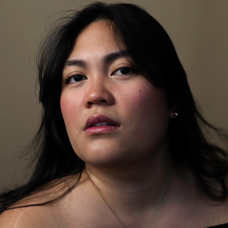 A woman with long dark hair and a neutral expression gazes slightly upward. She has light makeup and wears a subtle necklace. The background is plain and softly lit, highlighting her facial features and creating a warm, intimate portrait.