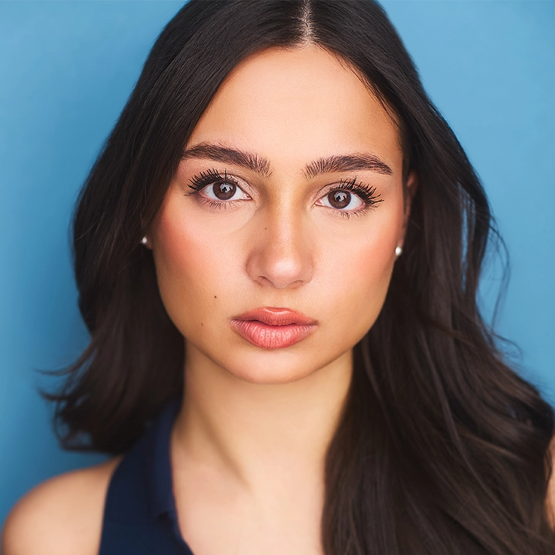 A young woman with long dark brown hair and brown eyes looks directly at the camera. Taylor Leccese has natural makeup, defined eyebrows, and wears a dark blue sleeveless top. The background is a solid light blue.