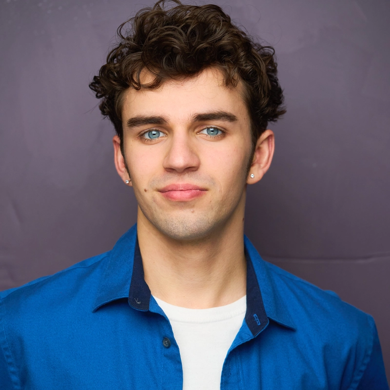 Carson Timmons, a young man with curly brown hair, blue eyes, and light skin, stands against a plain purple background. He wears small earrings, a blue collared shirt over a white T-shirt, and has a neutral, slight smile with a relaxed expression.