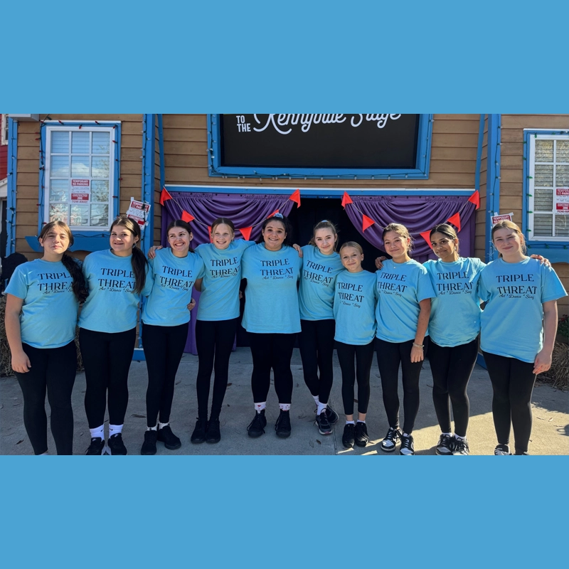 Ten girls stand side by side outside, all wearing matching light blue “TRIPLE THREAT” t-shirts from the Teen Triple Threat Program. They smile at the camera in front of a building with purple and red decorations and a sign reading “The Kenyonie Stage.”.