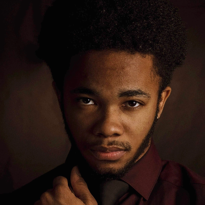 A young man with short, curly hair and a trimmed beard gazes directly at the camera. Channeling Carmelina Glynn’s signature style, he wears a dark burgundy shirt and adjusts his black tie under dramatic lighting against a shadowy background.