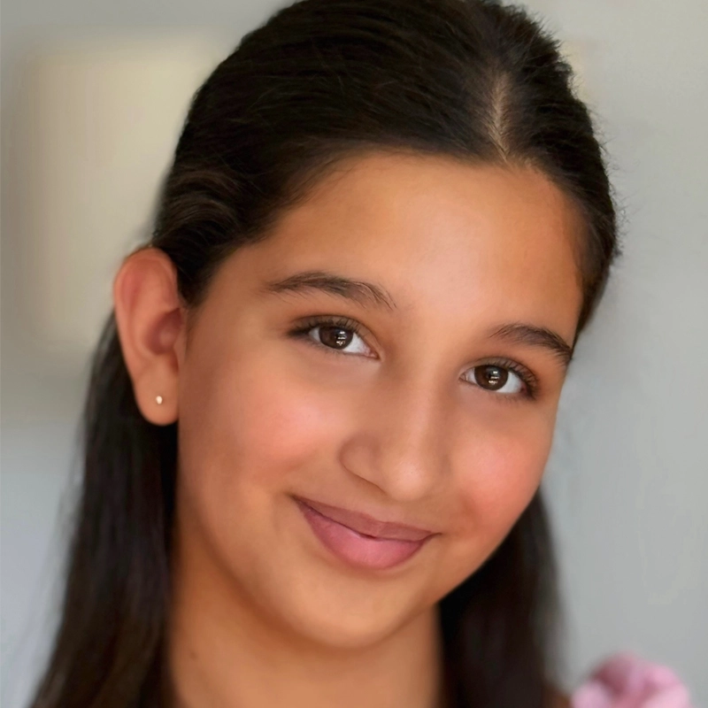 Victoria Underhill, a young person with long dark hair, brown eyes, and smooth skin, smiles gently at the camera. She wears a small earring and a light pink top; the softly blurred neutral background keeps focus on her face.