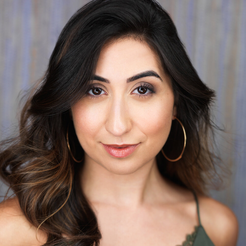 Krystal Hernandez, with long, wavy brown hair and large gold hoop earrings, poses facing forward. She has bold eyebrows, light makeup, and a subtle smile in a green spaghetti-strap top against a softly blurred blue-gray background.