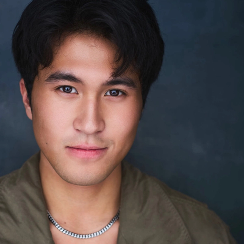 A young man with short, dark hair and smooth skin looks directly at the camera, smiling slightly. He wears a silver chain necklace and an olive-green jacket. The background is dark blue, softly blurred, drawing focus to his face.