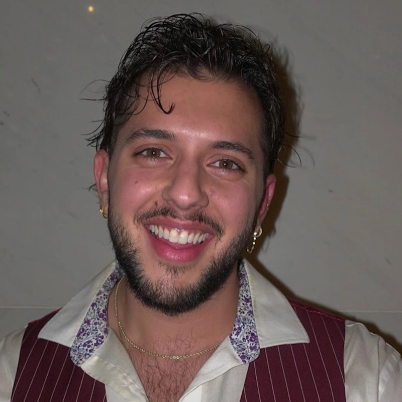 Casey Martin Klein, a young man with short brown hair and a beard, smiles at the camera. He wears a maroon pinstripe vest over a white collared shirt with floral inner lining, gold hoop earrings in both ears, and a gold necklace against a plain light-colored wall.