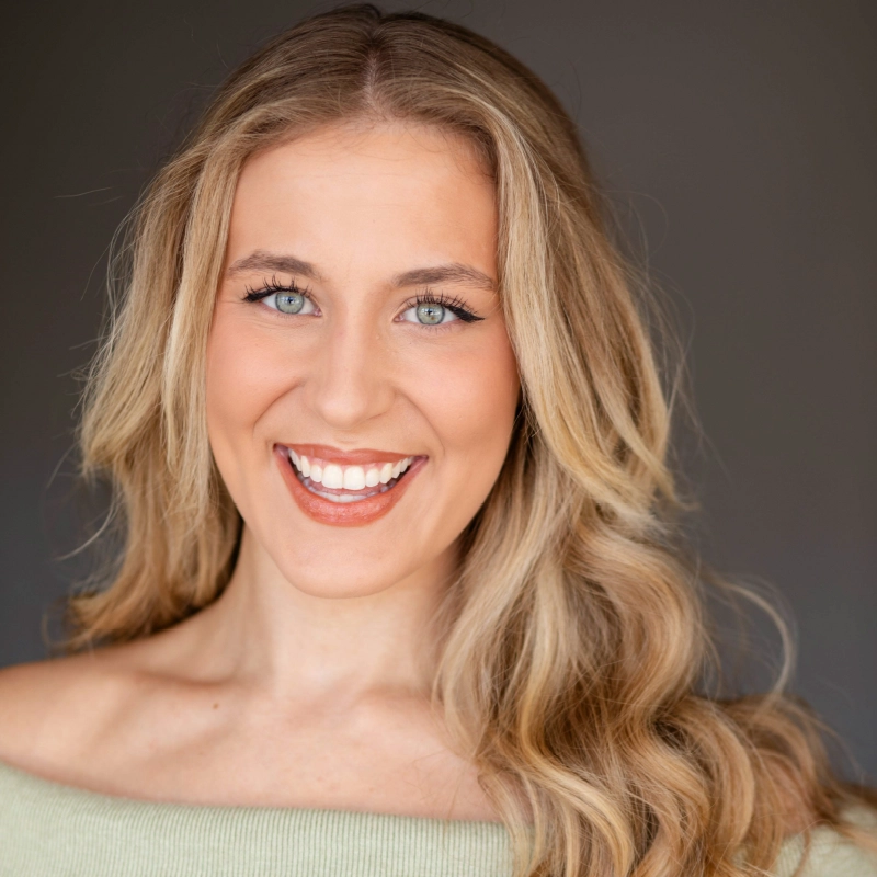 A young woman with long, wavy blonde hair smiles brightly at the camera. She has light skin, blue eyes, and wears natural makeup. She is dressed in a light green off-shoulder top and is set against a plain, neutral background.